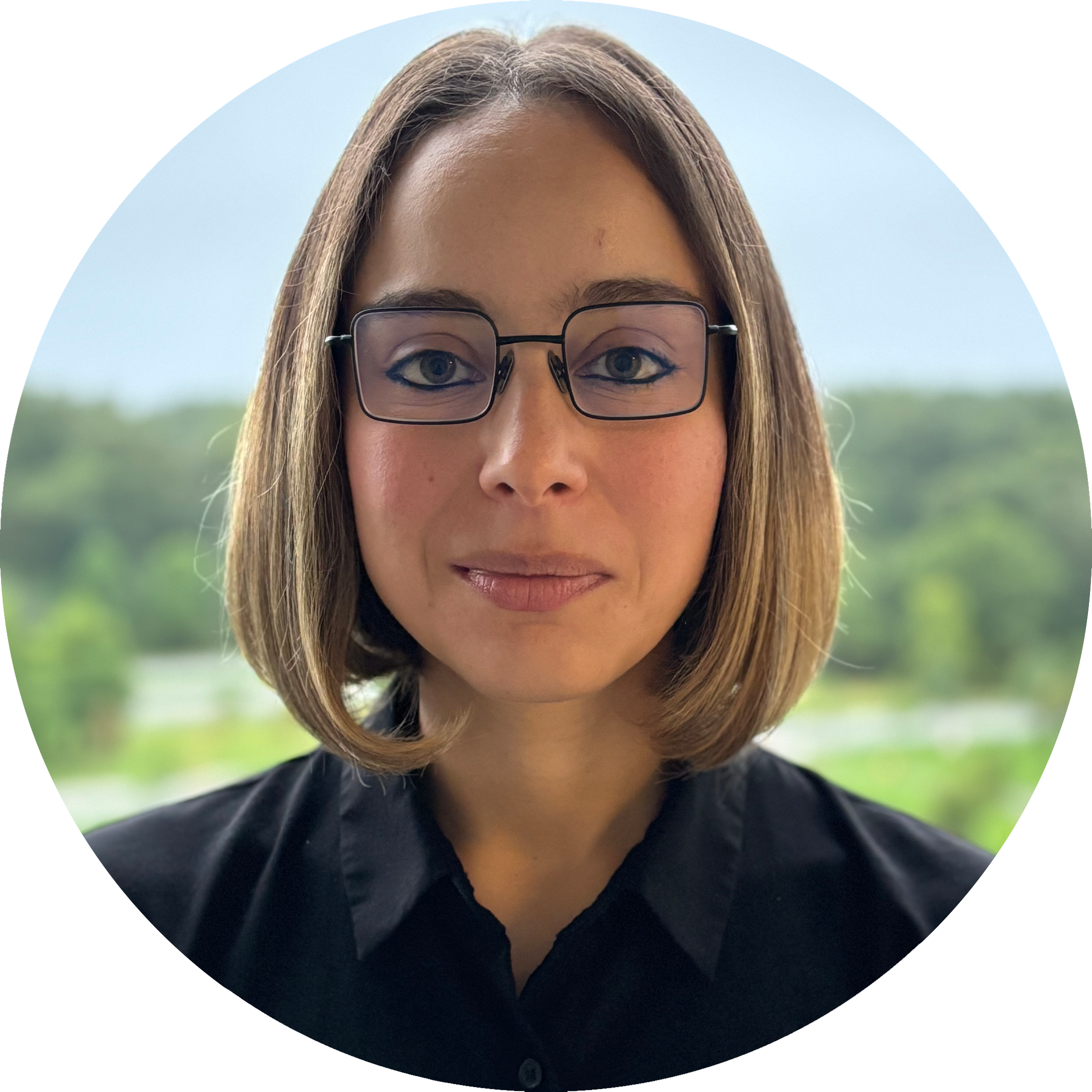 Headshot of Lina Marcella Carmona wearing glasses and a bob haircut, in a black shirt. Picture is set against a natural outdoor background