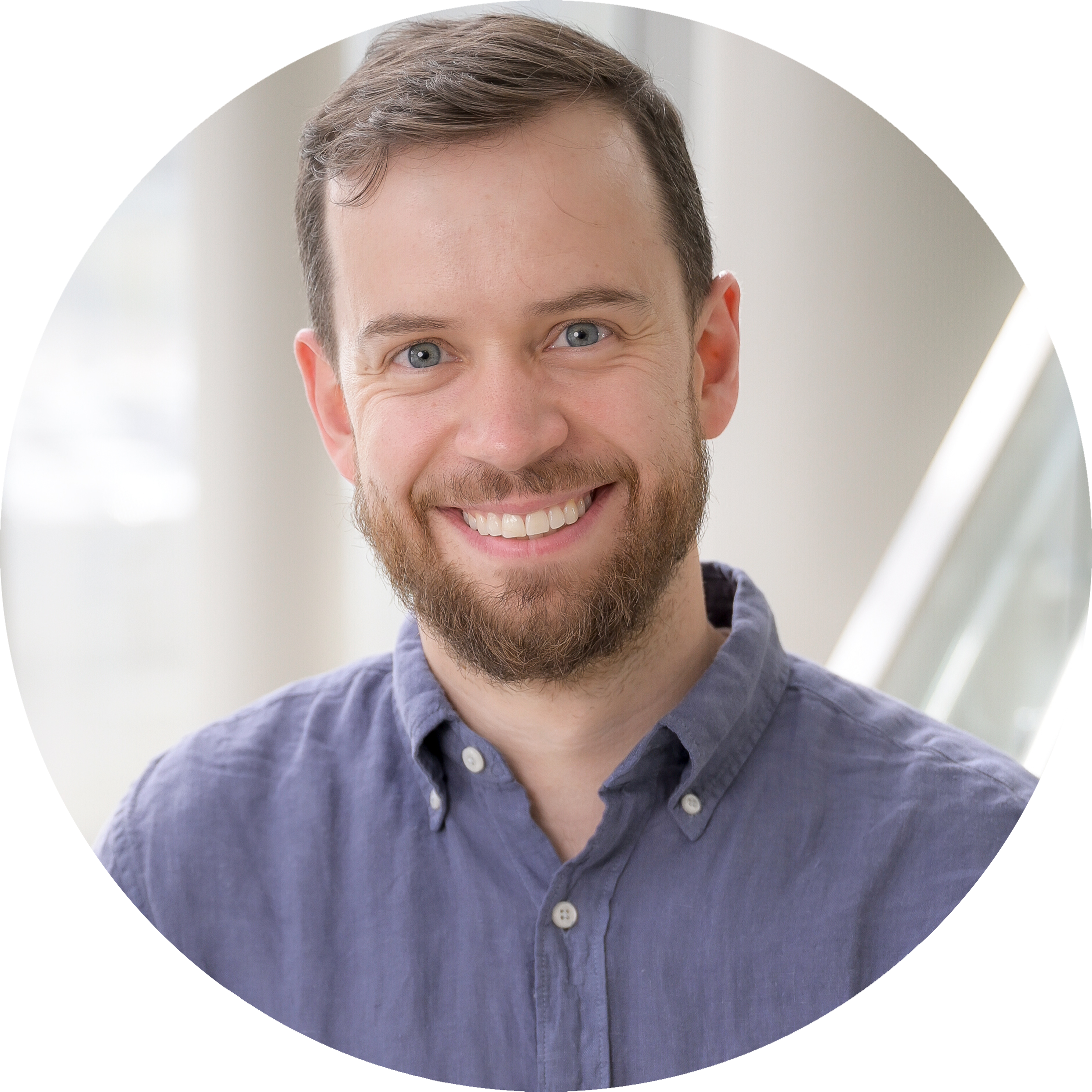 A headshot of Daniel Bondeson, wearing a smile, a beard, and a light blue, button-down shirt. The photo is set indoors with a white modern staircase railing in the background.