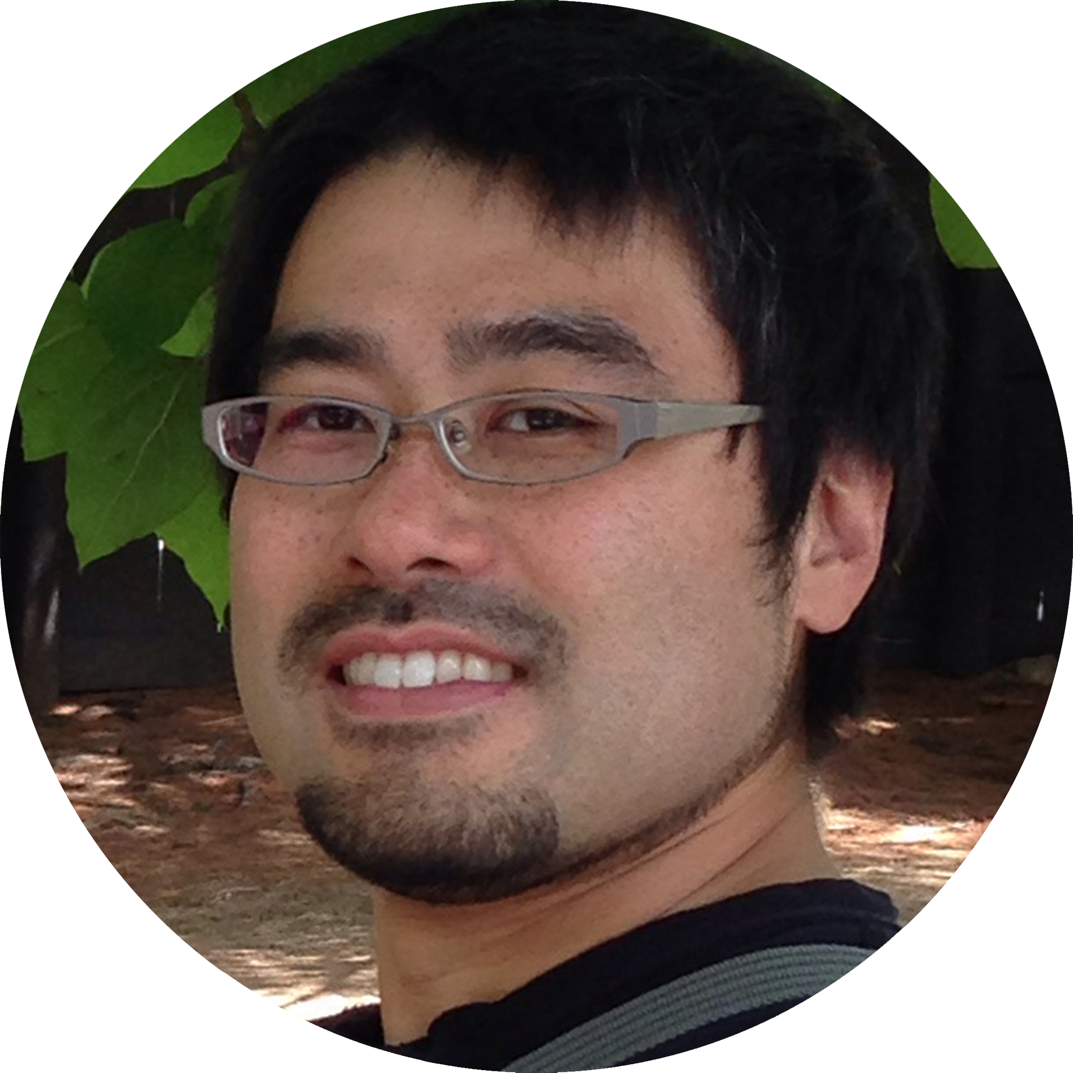 A selfie of Tadatoshi Sato wearing a broad smile in his glasses. He has a short dark hair and a goatee. The photo is set in front of large green leaves as a background.