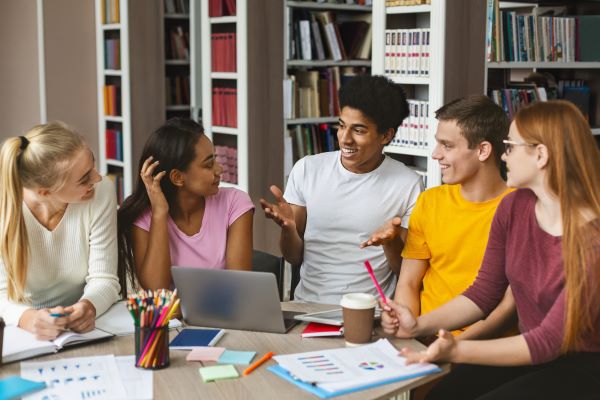 5 diverse teens smiling and chatting around a table with a laptop